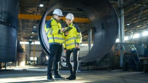 Male and Female Industrial Engineers in Hard Hats Discuss New Project while Using Tablet Computer. They're Making Calculated Engineering Decisions.They Work at the Heavy Industry Manufacturing Factory.