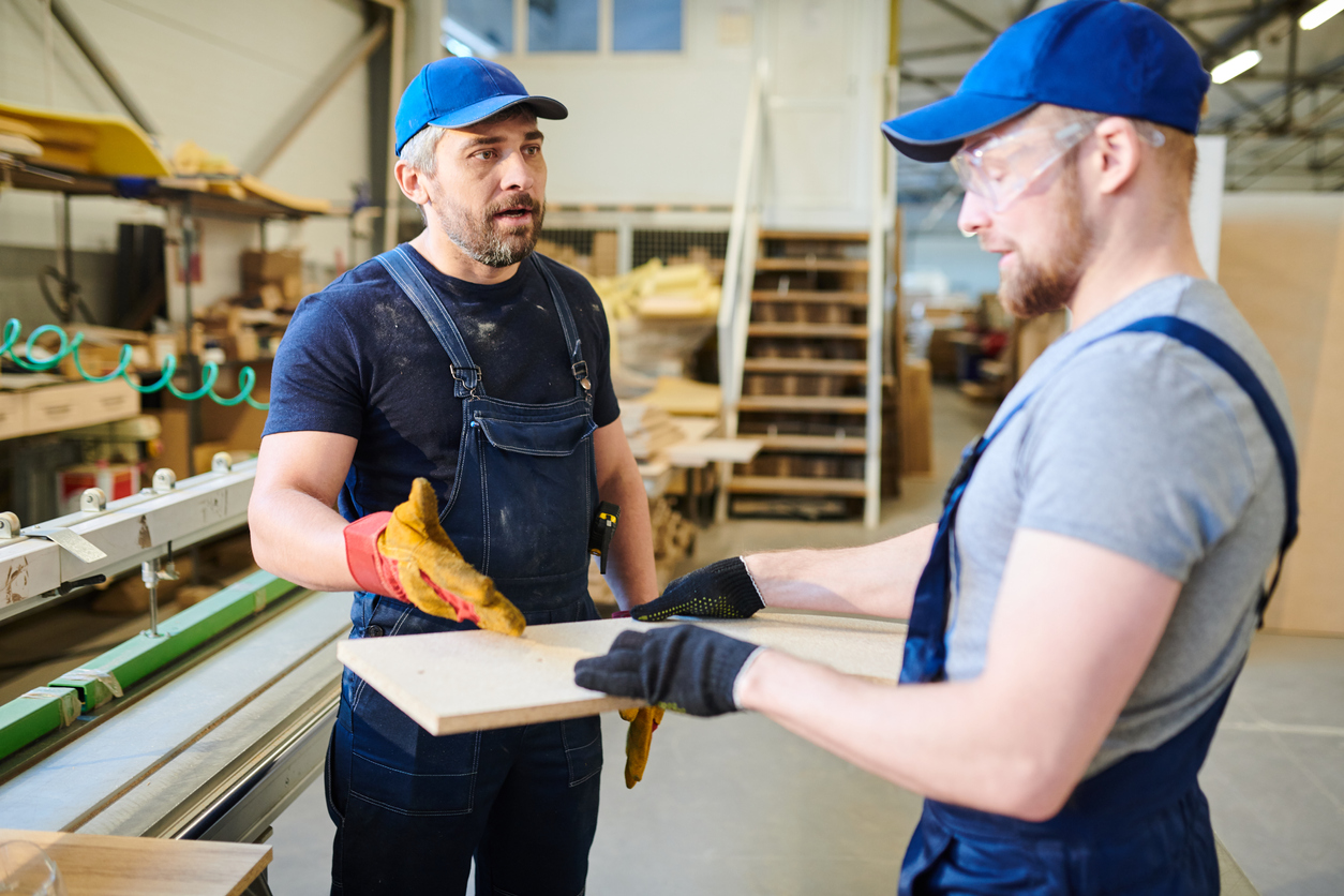 Serious displeased middle-aged bearded foreman in cap pointing at particleboard while explaining young worker how to make furniture