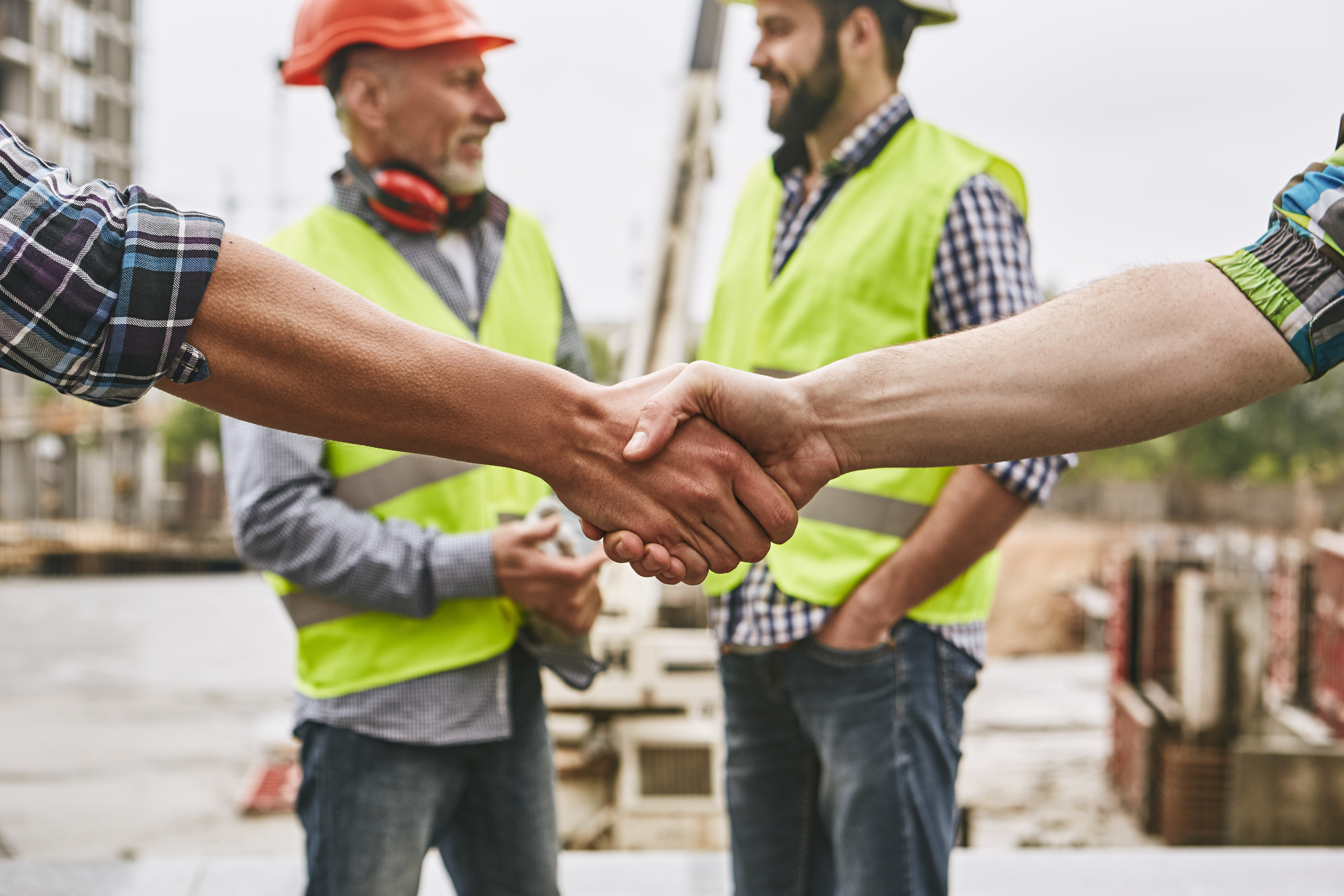 We did it! Close up photo of builders shaking hands against cheerful colleagues while working together at construction site. Team work.