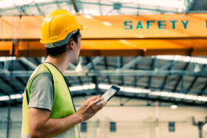 Young Asian male heavy industral worker using a digital tablet inside manufacturing and factory site with crane behind.